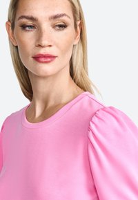 Top rosa con maniche a sbuffo e scollo rotondo, realizzato in tessuto morbido. Le maniche si raccolgono alla spalla per una silhouette strutturata.