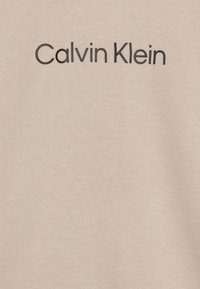 Bežová bavlnená tkanina s hladkou textúrou. Dizajn obsahuje text "Calvin Klein" v tučných čiernych písmenách cez hruď.