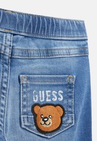 Denim-farkut vaaleansinisen pesun kanssa, joissa on takatasku, jossa on brodeerattu ruskeakarhun kuvio ja "GUESS" valkoisella kirjailtuna.