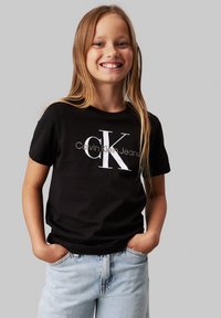 Melna kokvilnas t-krekls ar lielu baltu "CK" logotipu un tekstu "Calvin Klein Jeans". Saskaņots ar gaismas zilo džinsu šortiem.