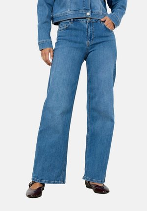 Person trägt hoch taillierte, weit geschnittene blaue Jeans und eine Jeansjacke, mit einer Hand in der Tasche und dunklen, glänzenden flachen Schuhen.