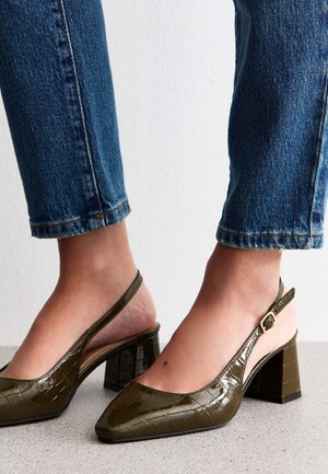 Escarpins à talon ouvert vert olive avec un bout pointu, finition brillante en motif crocodile et talon block. Associés à un jean bleu cropped.