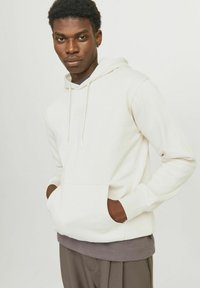Haut à capuche beige clair avec une poche à l'avant, un cordon de serrage, et des poignets côtelés. Porté sur une chemise gris foncé et associé à un pantalon marron.