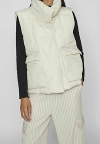 Vadderad, ärmlös väst i off-white med hög krage, som har två framfickor och lätta sömnadsdetaljer. Bärs över en svart topp.