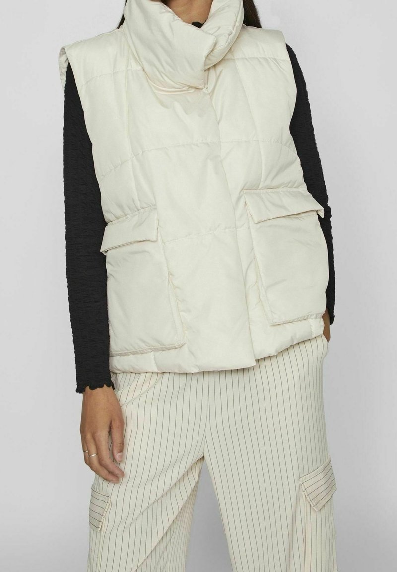 Vadderad, ärmlös väst i off-white med hög krage, som har två framfickor och lätta sömnadsdetaljer. Bärs över en svart topp.