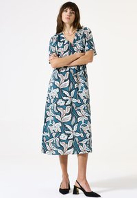 Türkise Blumenkleid in Midi-Länge mit kurzen Ärmeln, das eine taillierte Taille, filigrane weiße und rosafarbene Muster aufweist, sowie schwarze Pumps zum Hineinschlüpfen mit goldenen Akzenten.