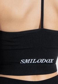 Top negro corto hecho de material acanalado con tirantes finos, que presenta el texto bordado en blanco "SMILODOX" en la banda inferior.