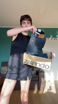 Oseba v črni obleki drži zalando škatlo in dviga par črnih kavbojk iz nje, stoji v sončni dnevni sobi.