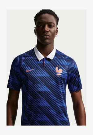 Uomo che indossa una maglia da calcio della Francia blu con colletto bianco, strisce diagonali, logo Nike e emblema del gallo a due stelle sul petto, sfondo neutro.