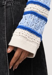 Χέρι με σκούρο τόνο δέρματος, μερικώς καλυμμένο από μανίκι πουλόβερ πλεκτού με κρέμα και ανοιχτό μπλε χρώμα και μπλε ρίγες, που ακουμπά πάνω σε μαύρο τζιν ύφασμα.