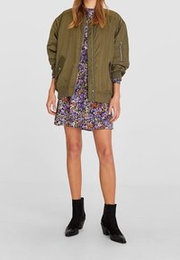 Mujer vestida con un vestido floral morado bajo una chaqueta bomber de gran tamaño en color verde oliva y botas negras hasta el tobillo.