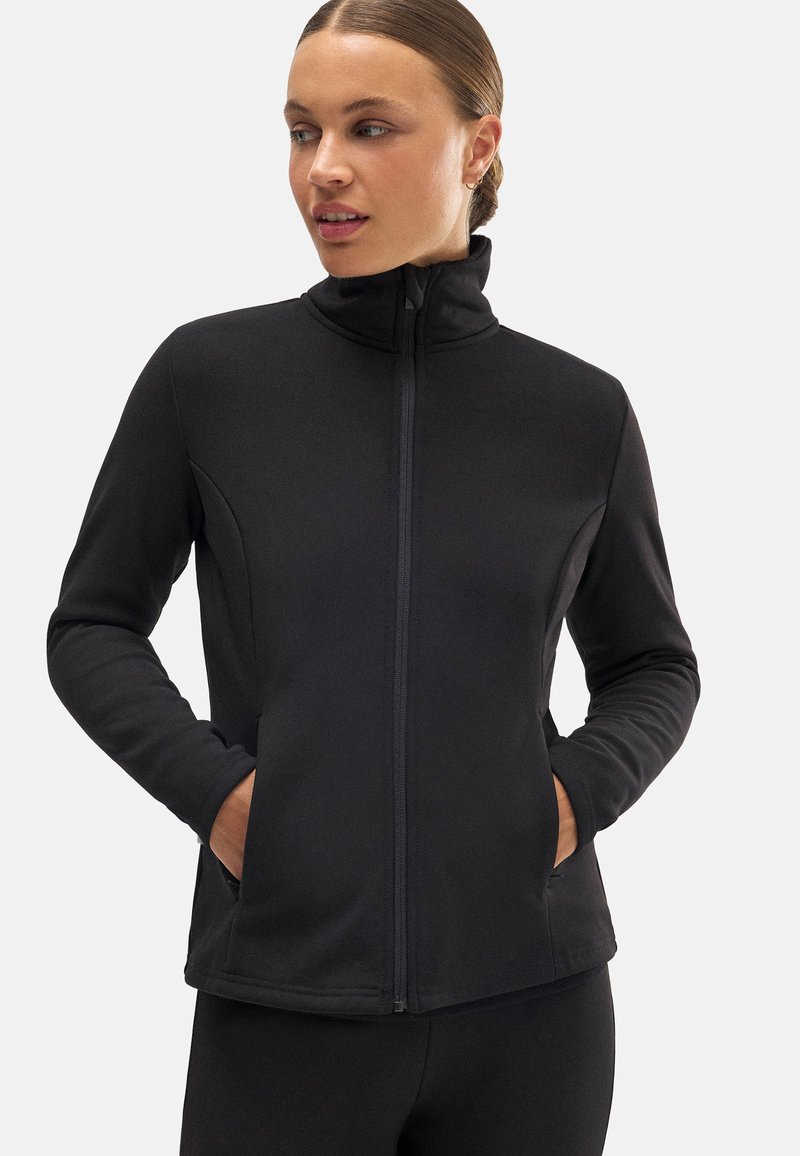 Chaqueta negra con cremallera y cuello alto, mangas largas y bolsillos laterales. Tejido suave con un diseño ajustado y mínimos detalles de costura.