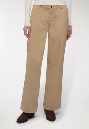 Pantaloni beige a gamba larga indossati con una maglietta bianca a maniche lunghe a costine e stivaletti neri con tacco, in piedi su uno sfondo bianco.
