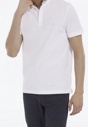 Homme portant un polo blanc uni à manches courtes et un pantalon foncé, une main près du cou et l'autre le long du corps.