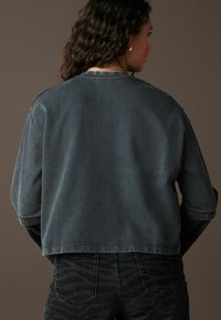 Mørkegrå denimjakke med løstsiddende pasform, der har et cropped design og opbrettede ærmer. Detaljeret syning langs kanterne.