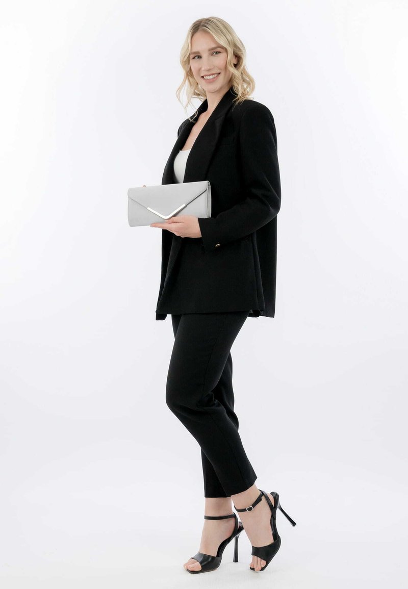 Černý blazer přes bílý top, černé zúžené kalhoty, stříbrný psaníčko s V-tvarovaným kovovým detailem. Černé sandály na vysokém podpatku s řemínky kolem kotníků.