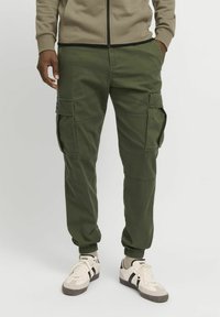 Mand iført olivengrønne cargobukser med sidelommer og beige sneakers med sorte striber, stående mod en ensfarvet baggrund.