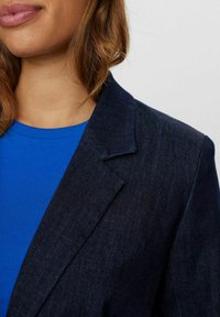 Navy blue denim blazer with sharp lapels, textured fabric, and stitching detail, paired with a ribbed blue top.