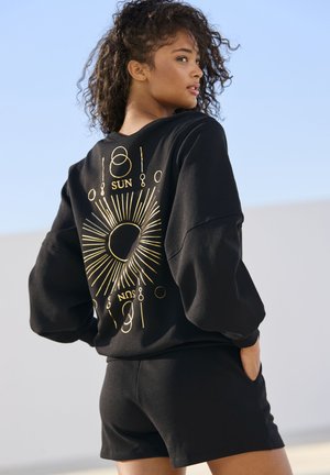 Black sweatshirt with gold sun and moon graphic on the back, paired with black shorts. Fabric appears soft with a relaxed fit.