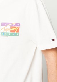 Camiseta de algodón blanca con un colorido logo de "TOMMY JEANS" que presenta elementos en púrpura, naranja y verde. Pequeño acento de marca en la manga.