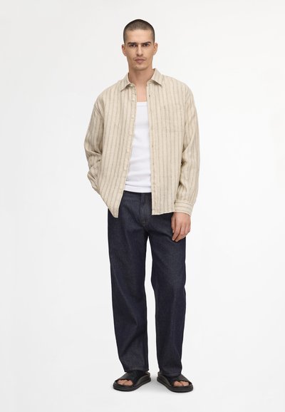 Man standing wearing beige striped button-up shirt over white tank top, dark blue jeans, and black open-toe sandals against white background.
