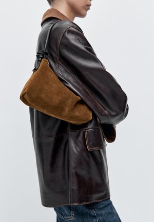 Braune Leder- und Wildleder-Schultertasche, leicht abgerundete Form, mit verstellbarem Riemen und dezenten Texturen, getragen zu einer braunen Lederjacke.