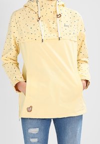 Gul anorak med ett tryckt mönster av små färgade trianglar, justerbar huva med dragsnören och framficka. Har en brun logotypapplikation.