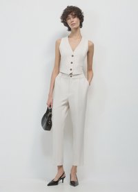 Weißes ärmelloses Oberteil mit Knopfverschluss, kombiniert mit einer schlank sitzenden Hose. Das Model hält eine schwarze Handtasche und trägt schwarze Slingback-Absätze.
