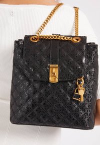 Mochila sintética negra con patrón de logo en relieve, herrajes en tono dorado, una correa de cadena y un charm de candado decorativo. Textura suave y brillante.