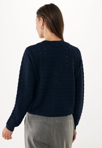 Námořnický modrý pletený svetr s vlnovým vzorem a dlouhými rukávy. Hribované manžety a výstřih, spárované se šedými oblekovými kalhotami.