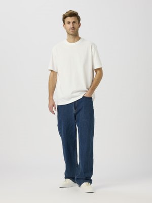White cotton t-shirt paired with loose-fitting dark blue denim jeans. Model wears white sneakers, with a relaxed stance against a plain background.