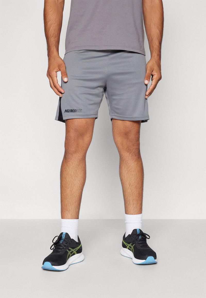 Lichtgrijze sportshorts met zwarte accenten, logo op de linkerdij, gecombineerd met zwarte sneakers en witte enkelsokken.