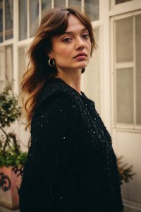 Noore naise laineliste pruunide juustega, kes kannab rõngaskõrvarõngaid ja musta sädelevat jopet, seisab väljas akende ja taimede lähedal.