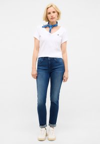 Tricou V cu guler din bumbac alb, eșarfă albastră cu model legată la gât, blugi albaștri închis și adidași albi. Modelul stă în fața unui fundal deschis.