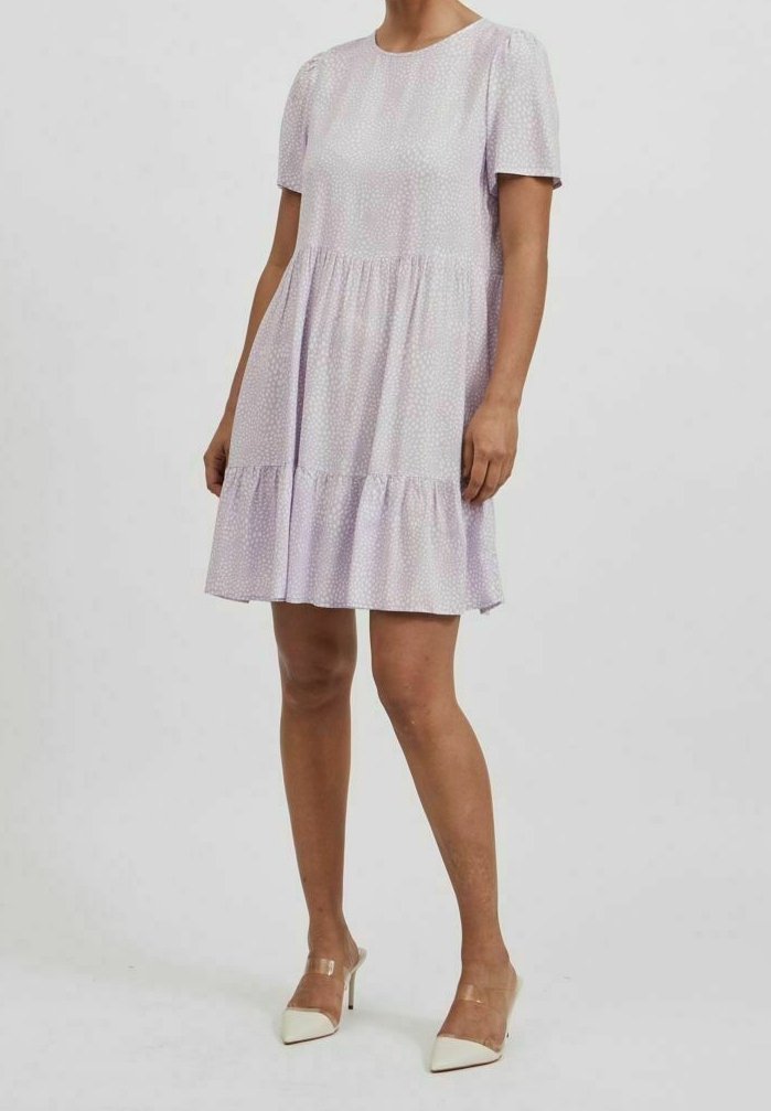 Žena v krátkých sl sleeves světle fialových šatech s vrstveným designem a malým tečkovaným vzorem, doplněná bílými špičatými podpatky s průhlednými pásky.
