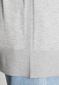 Světle šedý pletený svetr s vroubkovaným lemem a bočním detailem; má měkkou strukturu a volný střih, kombinovaný s modrými džíny.