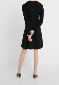 Robe en tricot côtelé noire avec des manches longues, un buste ajusté et une jupe évasée. Le dos présente un col rond et est assorti à des chaussures à talons noires.