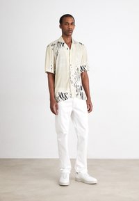 Short-sleeve shirt in light beige with black and cream abstract patterns, paired with white pants and white sneakers, standing on a neutral background.