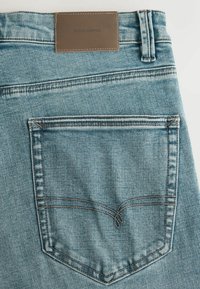 Világoskék farmernadrág, hátul barna kontrasztos varrással ellátott zsebbel és a derékon "DENIM SUPPLY" feliratú bőr címkével.