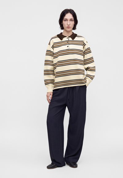 Femme aux cheveux courts et foncés portant un polo à manches longues rayé crème et marron ainsi qu'un pantalon ample bleu marine foncé, debout devant un arrière-plan uni.