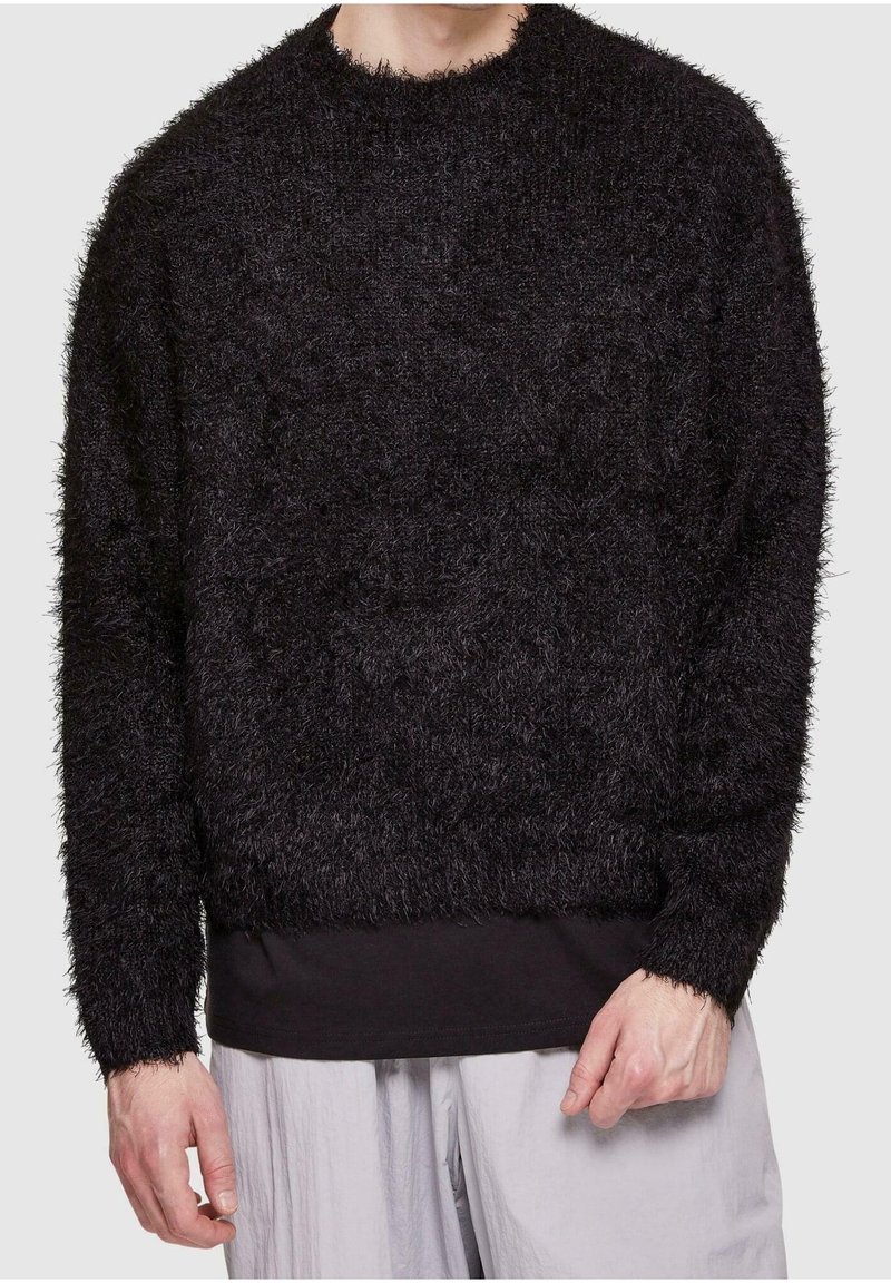 Homme portant un pull noir à manches longues et en matière duveteuse par-dessus une chemise noire, avec un pantalon gris clair, debout les mains détendues le long du corps.