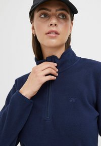 Pull-over en polaire bleu marine avec un col montant, design à demi-fermeture éclair et logo brodé. Texture douce et manches longues avec poignets.