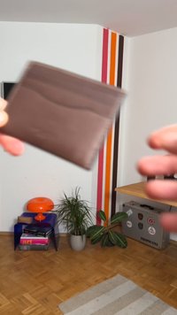 Brūna ādas maks ar plānu dizainu, kam raksturīga gluda tekstūra un smalkas locījuma līnijas, turēts starp divām rokām uz neitrāla fona.