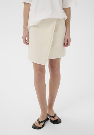 Naine, kes kannab kreemjat tekstuuriga mähissahtlit, valget lühikeste varrukatega topi ja musti ruudukujulisi varbavahe-sandaale, seisab valgel taustal.