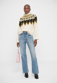 Maglione bianco a collo alto lavorato a maglia con motivi geometrici neri, gialli e grigi, abbinato a jeans svasati blu chiaro e una borsa rosa.