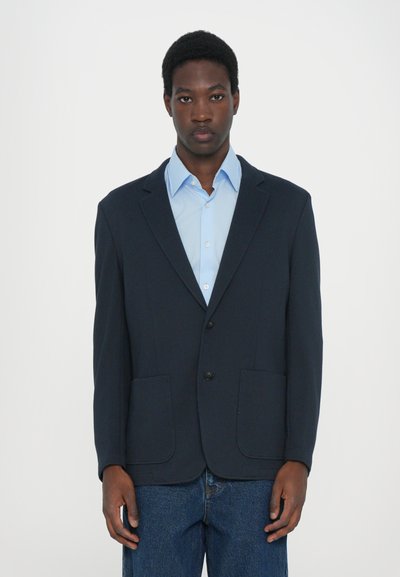 Blazer navy avec des revers crantés, deux poches avant et fermeture à boutons, assorti à une chemise bleu clair et un pantalon en denim. Coupe structurée.