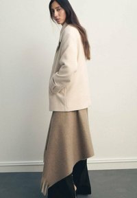 Cappotto in misto lana beige con tasche laterali, abbinato a una lunga gonna taupe asimmetrica con bordi frangiati e accenti neri.