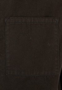 Brązowa bawełniana tkanina z kwadratową nakładką kieszeni z widocznym przeszyciem. Faktura wydaje się gładka, z niewielkimi niedoskonałościami materiału.