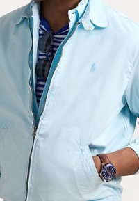 Ljusblå zip-up jacka med krage, med framfickor och en broderad logotyp, bärs över en randig skjorta i marinblått och vitt.