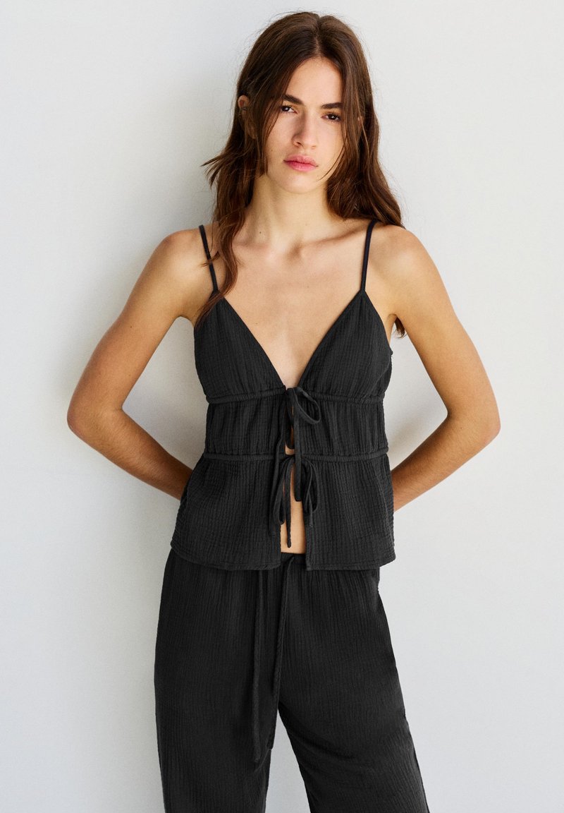 Jeune femme aux longs cheveux bruns se tient debout, les mains dans le dos, portant un haut noir sans manches avec des liens à l'avant et un pantalon ample assorti.